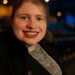 A smiling woman with light brown hair and red lipstick, wearing a black jacket over a gray sweater, is shown in a softly lit close-up portrait. The background is gently blurred with warm and cool tones, creating a calm and inviting atmosphere.