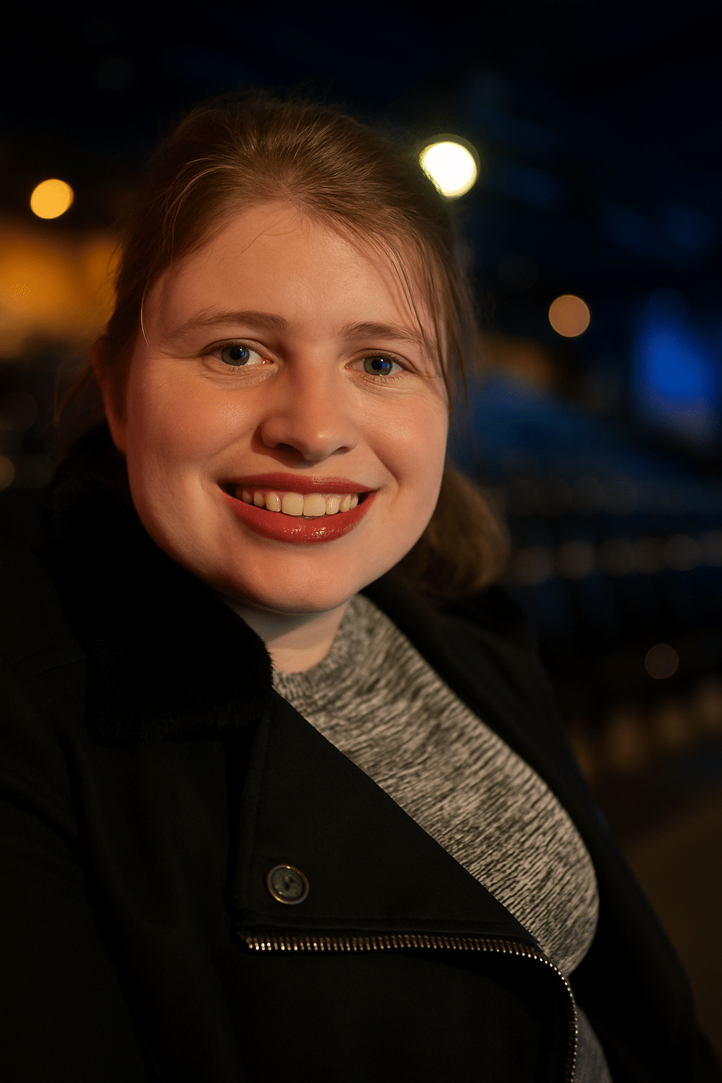 A smiling woman with light brown hair and red lipstick, wearing a black jacket over a gray sweater, is shown in a softly lit close-up portrait. The background is gently blurred with warm and cool tones, creating a calm and inviting atmosphere.